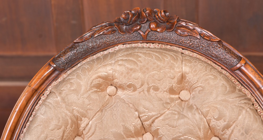 A Victorian carved walnut nursing chair with a button-tufted oval backrest, champagne fabric upholstery, carved cabriole legs, and ceramic castors. View XI