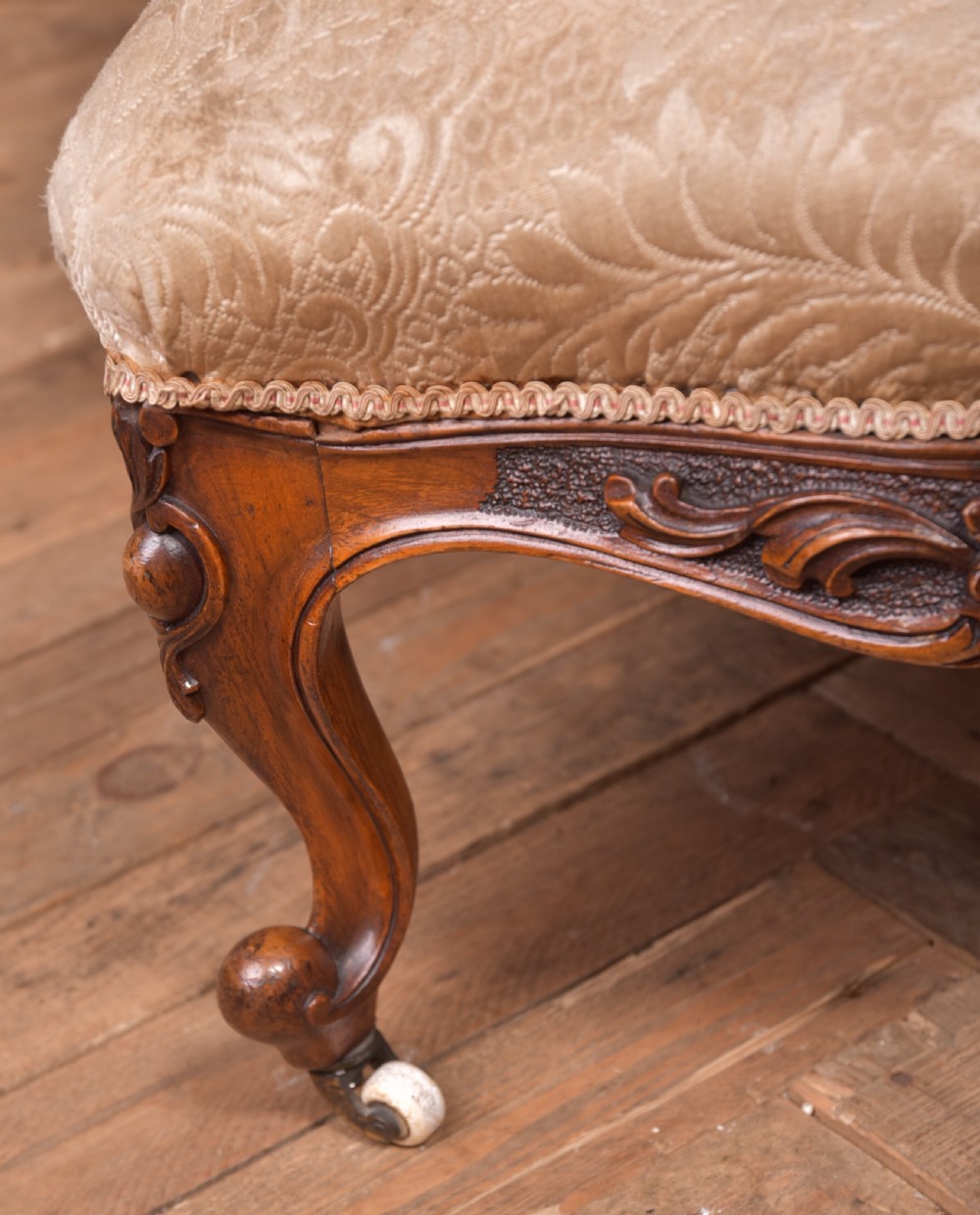 A Victorian carved walnut nursing chair with a button-tufted oval backrest, champagne fabric upholstery, carved cabriole legs, and ceramic castors. View XII