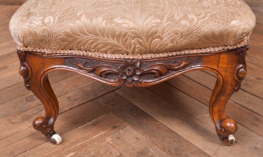 A Victorian carved walnut nursing chair with a button-tufted oval backrest, champagne fabric upholstery, carved cabriole legs, and ceramic castors. View XIII