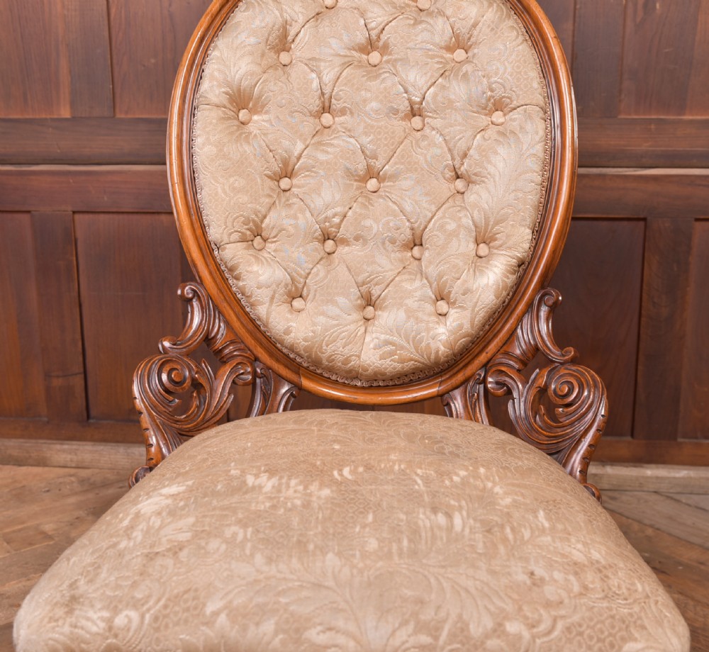 A Victorian carved walnut nursing chair with a button-tufted oval backrest, champagne fabric upholstery, carved cabriole legs, and ceramic castors. View XIV