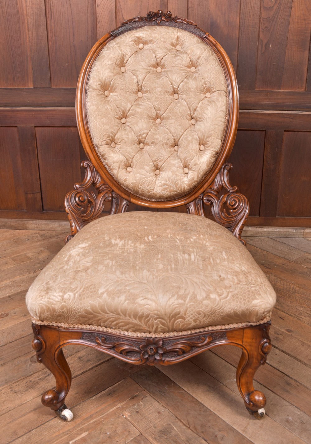 A Victorian carved walnut nursing chair with a button-tufted oval backrest, champagne fabric upholstery, carved cabriole legs, and ceramic castors. View XV