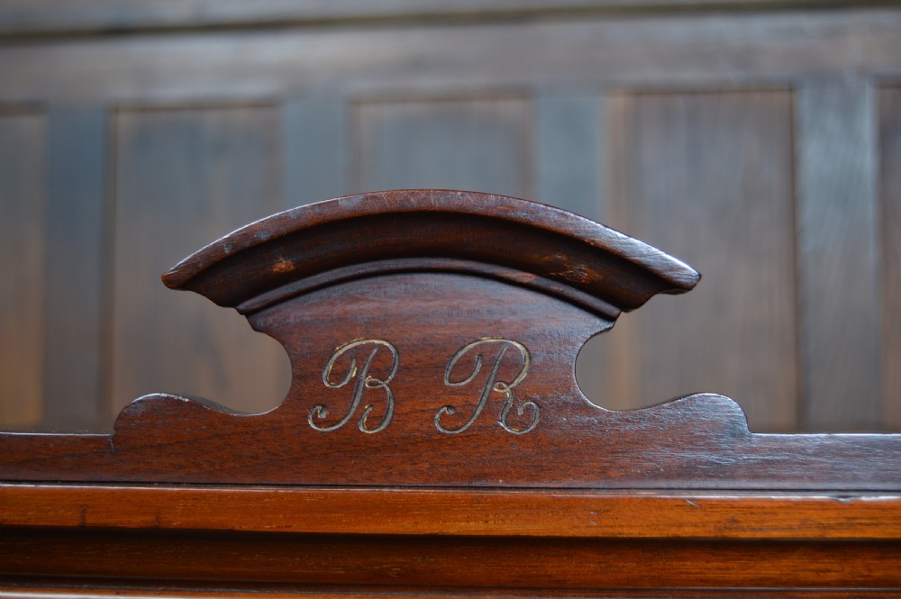 An Edwardian miniature mahogany wardrobe featuring mirrored twin doors, three lower drawers with brass drop handles, and an arched crest inscribed with initials. View IX