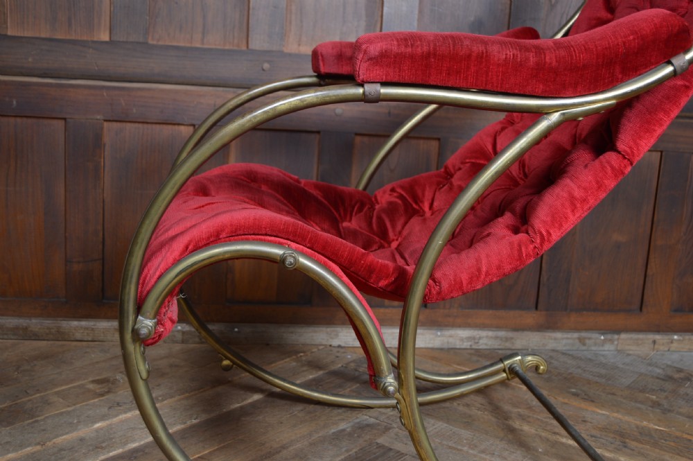 An Edwardian rocking chair featuring a tubular brass curved frame with button-tufted red velvet upholstery and padded armrests. View X
