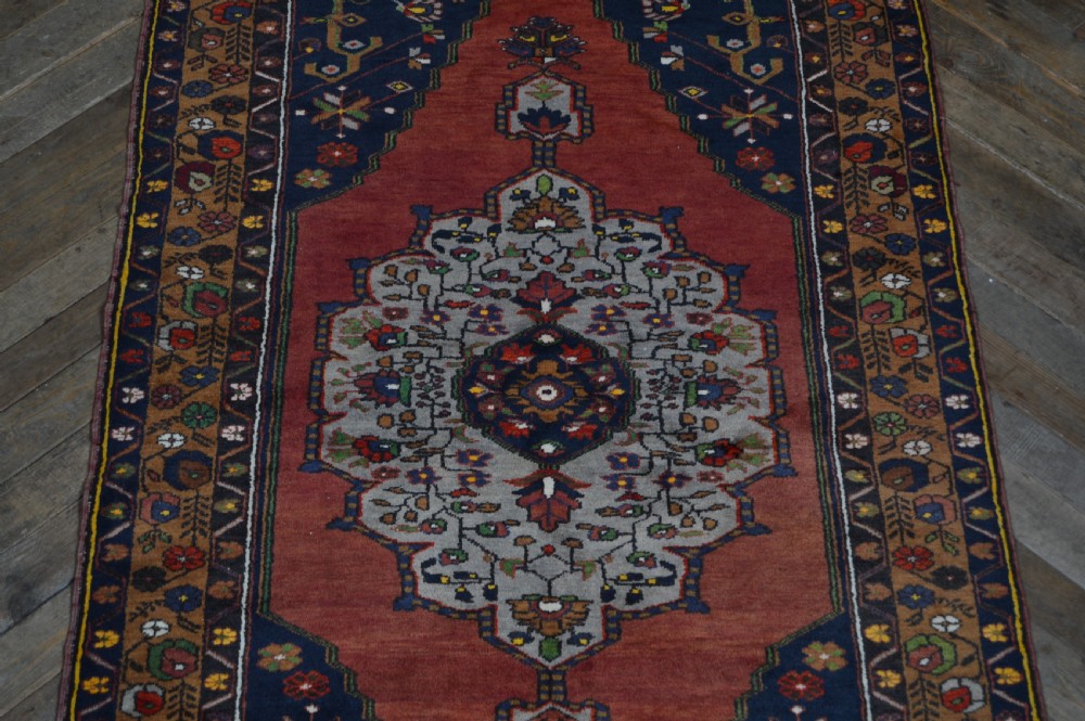A long hand-knotted Turkish rug runner featuring a central light grey and blue geometric medallion on a terracotta red field, framed by dark navy spandrels and a multi-band decorative border. View IV