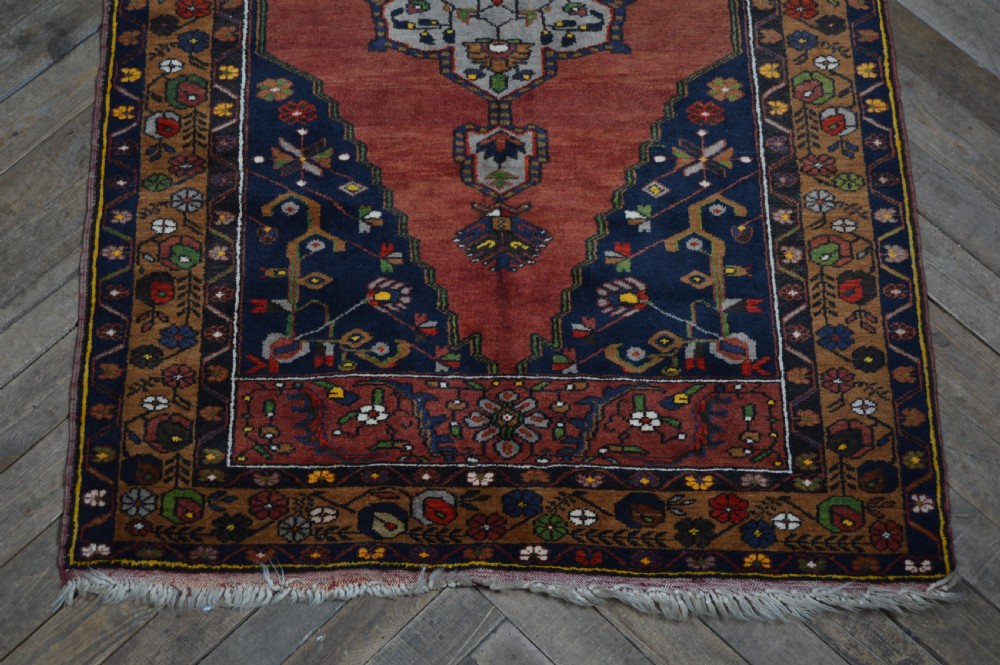 A long hand-knotted Turkish rug runner featuring a central light grey and blue geometric medallion on a terracotta red field, framed by dark navy spandrels and a multi-band decorative border. View V