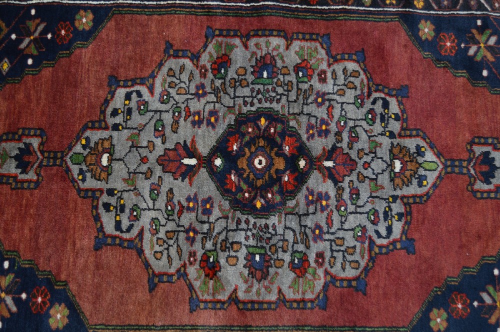 A long hand-knotted Turkish rug runner featuring a central light grey and blue geometric medallion on a terracotta red field, framed by dark navy spandrels and a multi-band decorative border. View VII