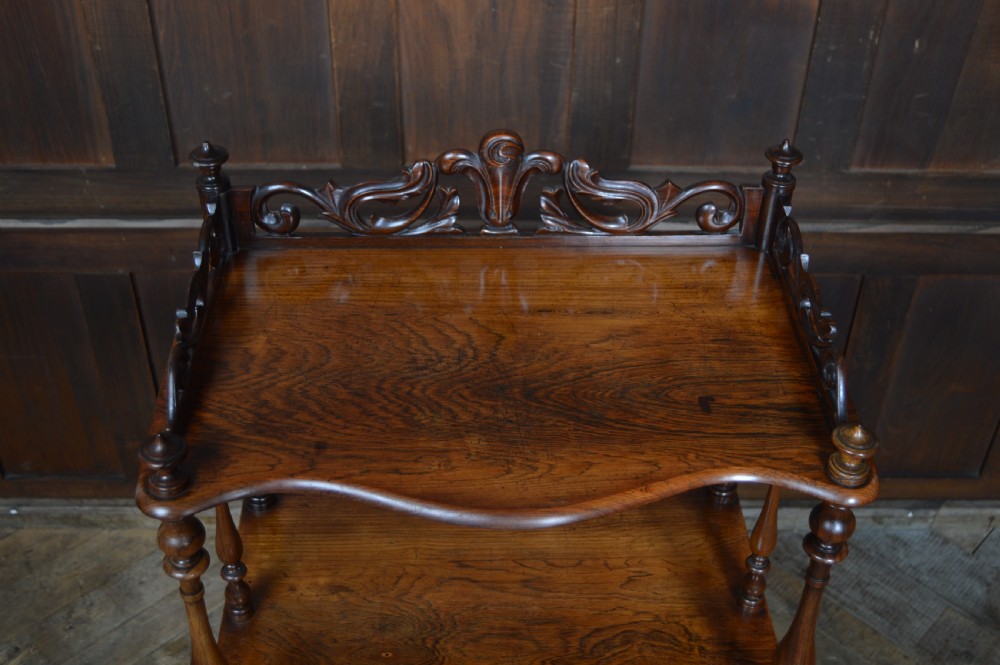 A three-tier Victorian rosewood whatnot featuring turned spindle supports, a carved cresting gallery back, shaped shelf edges, and brass castors. View V