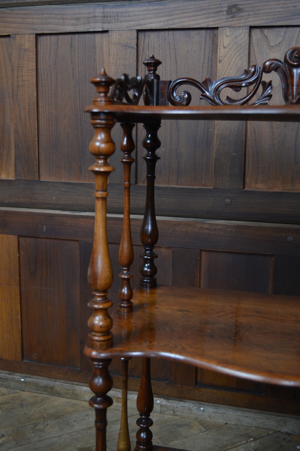 A three-tier Victorian rosewood whatnot featuring turned spindle supports, a carved cresting gallery back, shaped shelf edges, and brass castors. View X