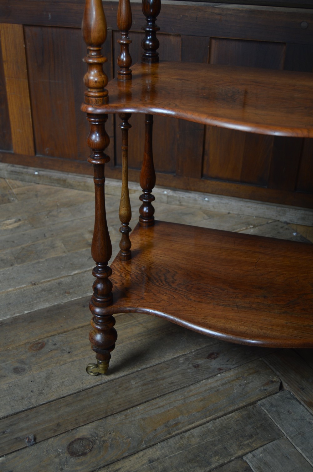 A three-tier Victorian rosewood whatnot featuring turned spindle supports, a carved cresting gallery back, shaped shelf edges, and brass castors. View XI