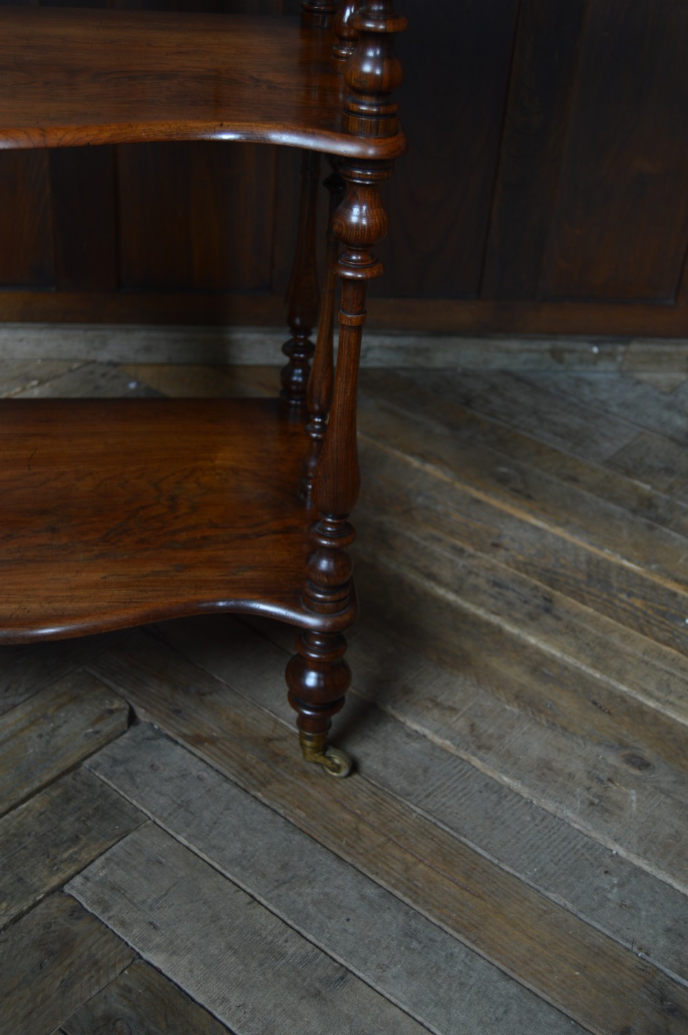 A three-tier Victorian rosewood whatnot featuring turned spindle supports, a carved cresting gallery back, shaped shelf edges, and brass castors. View XII