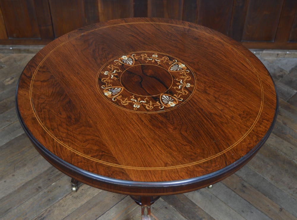 A circular Edwardian rosewood centre table featuring central floral marquetry inlay, edge stringing, and a turned pedestal with four splayed legs on brass castors. View III