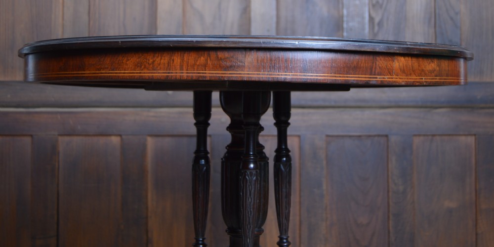 A circular Edwardian rosewood centre table featuring central floral marquetry inlay, edge stringing, and a turned pedestal with four splayed legs on brass castors. View VII