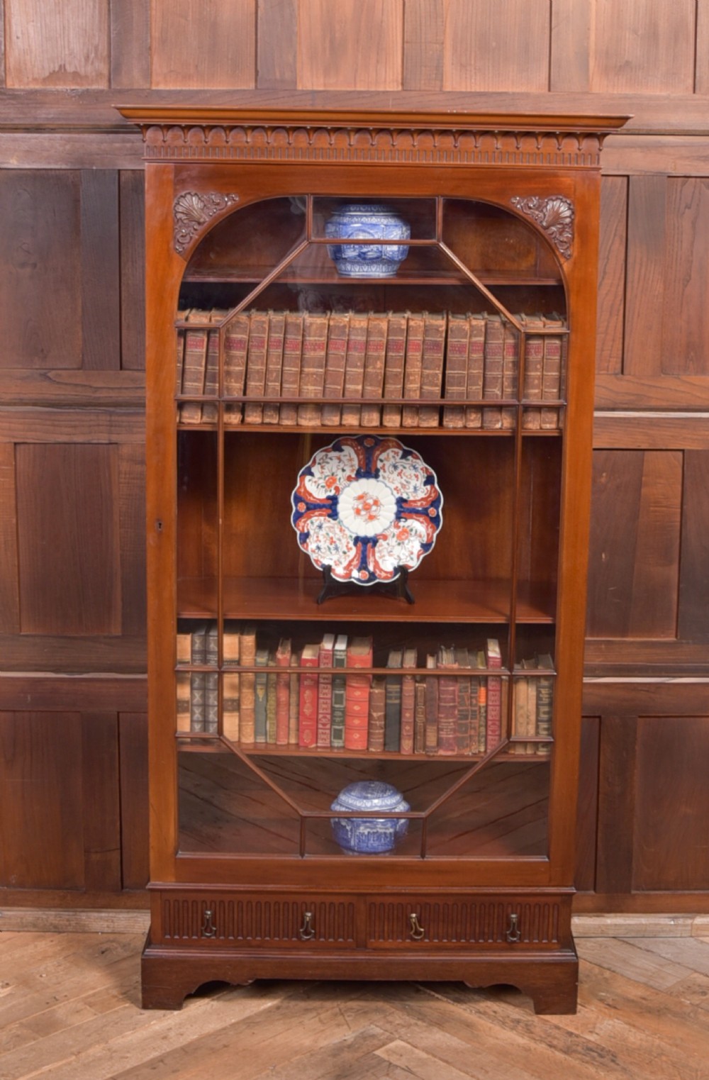 An Edwardian mahogany glazed bookcase featuring geometric astragal glazing, carved corner spandrels, and bottom drawers with drop handles.