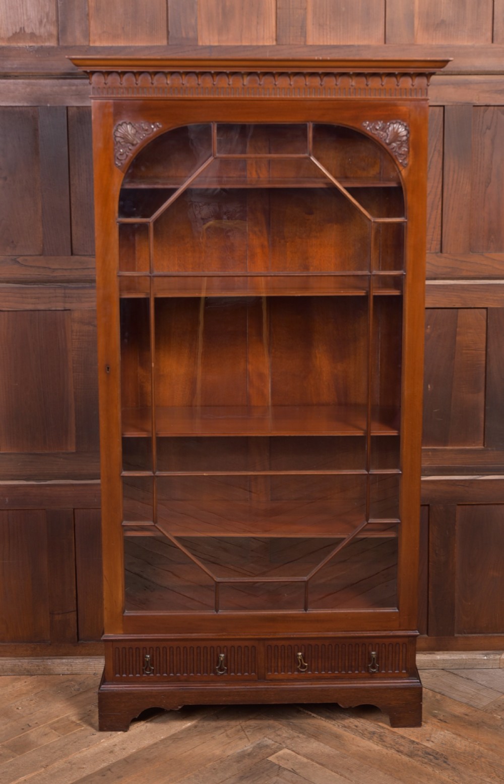 An Edwardian mahogany glazed bookcase featuring geometric astragal glazing, carved corner spandrels, and bottom drawers with drop handles. View II