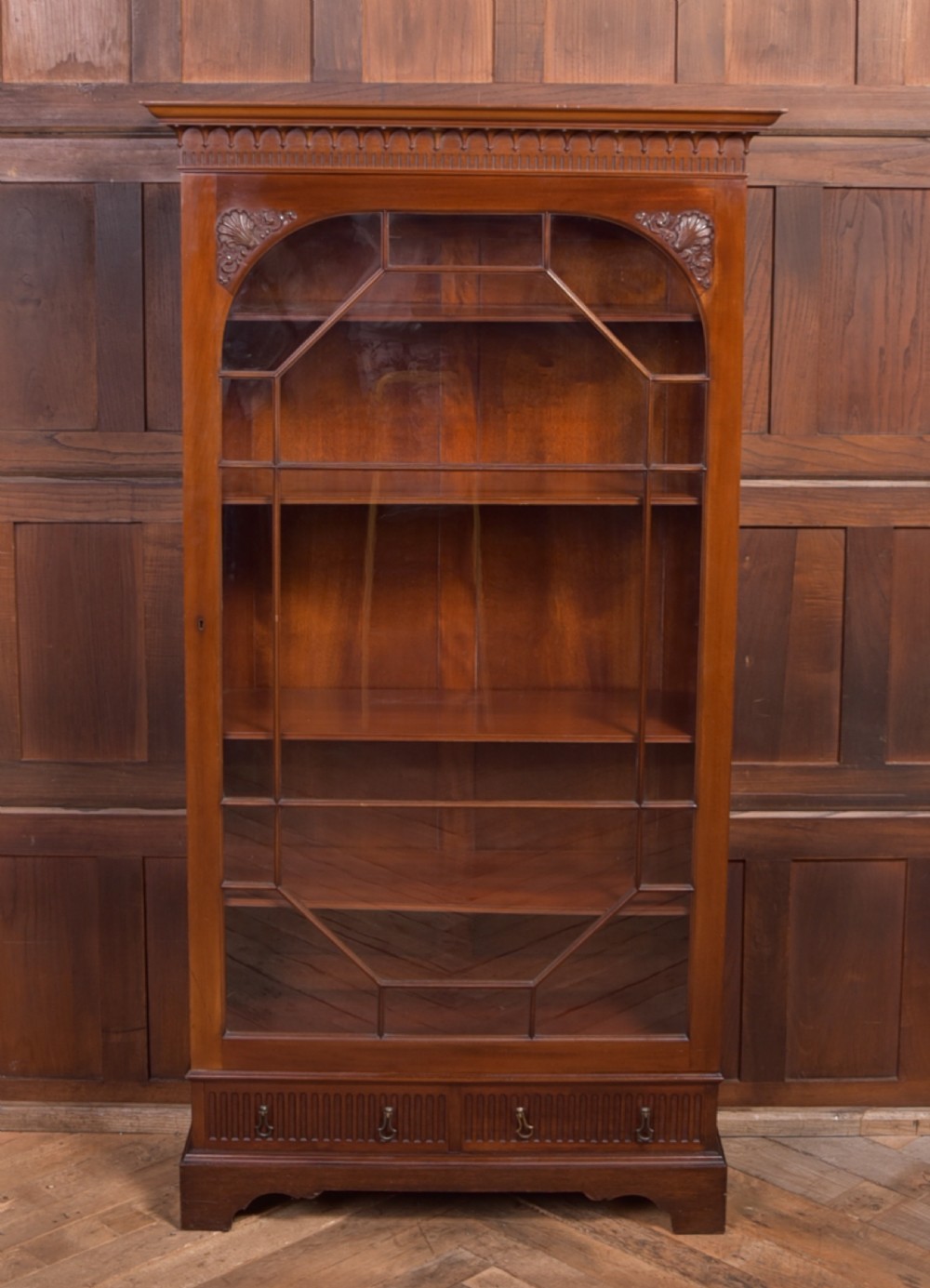 An Edwardian mahogany glazed bookcase featuring geometric astragal glazing, carved corner spandrels, and bottom drawers with drop handles. View VII
