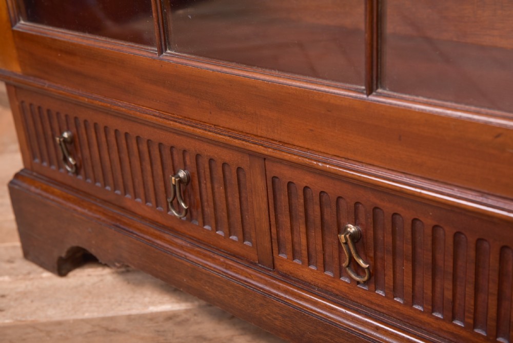 An Edwardian mahogany glazed bookcase featuring geometric astragal glazing, carved corner spandrels, and bottom drawers with drop handles. View X