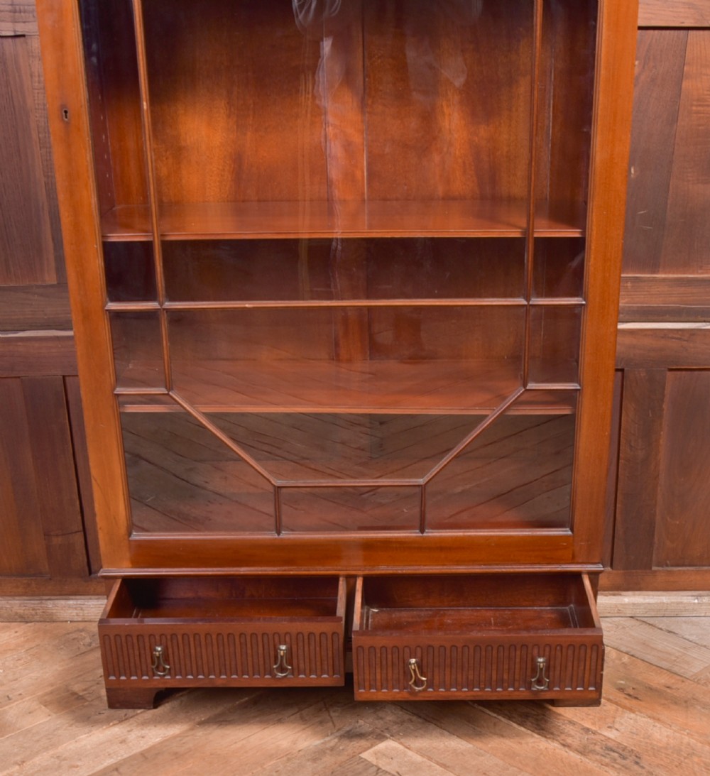 An Edwardian mahogany glazed bookcase featuring geometric astragal glazing, carved corner spandrels, and bottom drawers with drop handles. View XVII