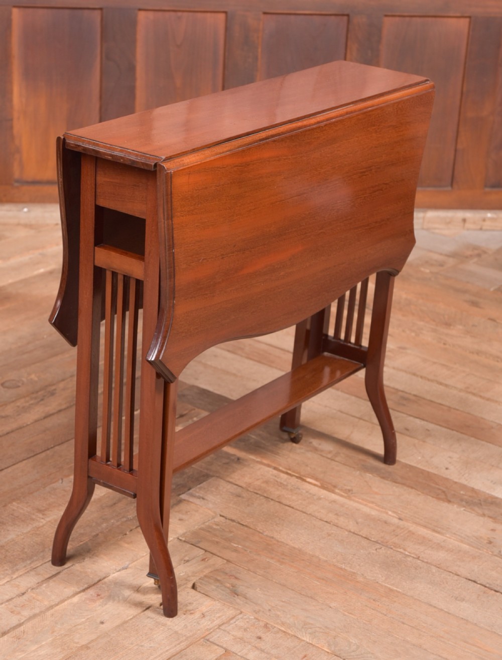An Edwardian mahogany Sutherland side table shown in a folded state with deep drop leaves, slatted end supports, and curved legs with brass castors.