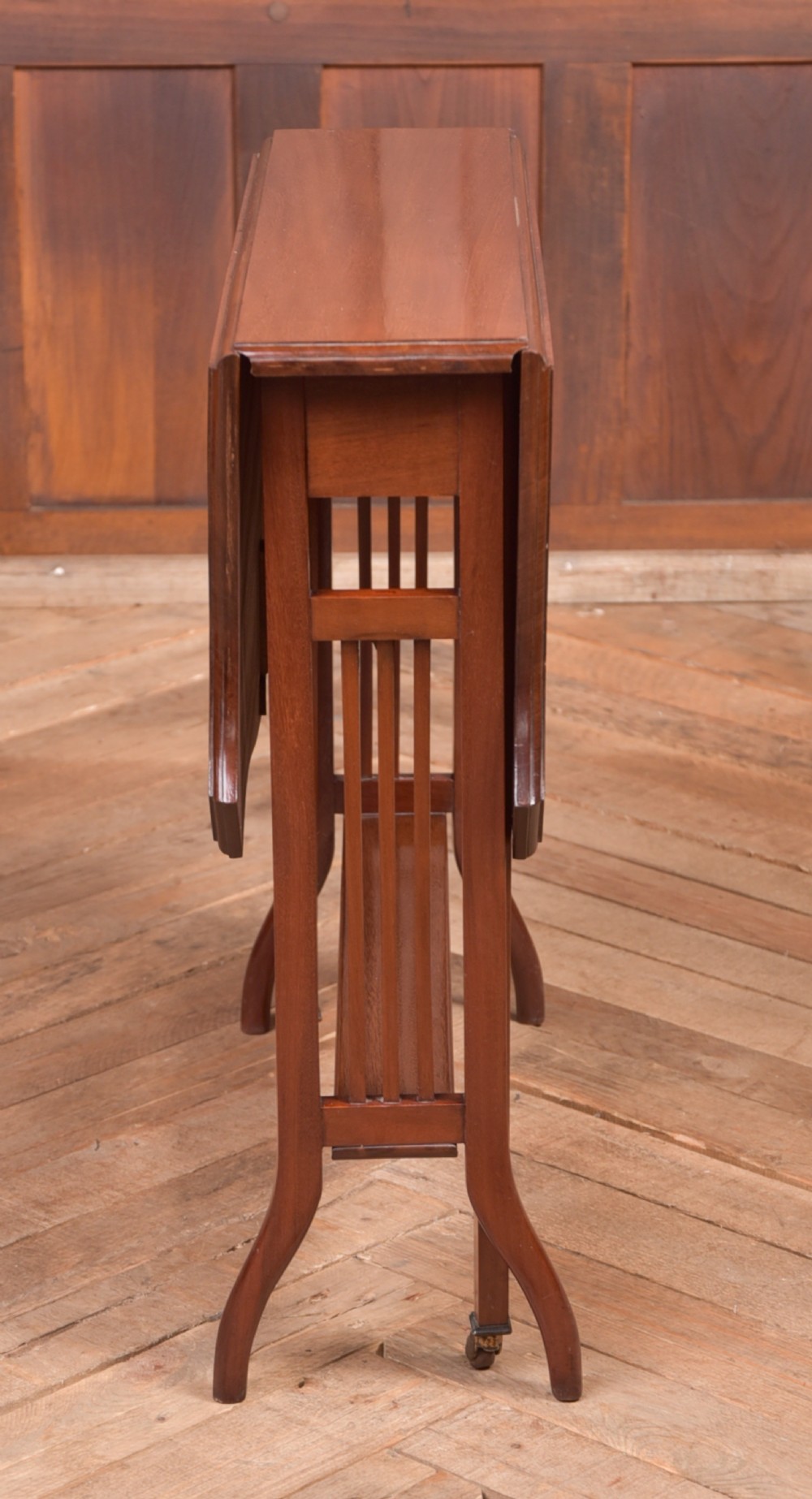 An Edwardian mahogany Sutherland side table shown in a folded state with deep drop leaves, slatted end supports, and curved legs with brass castors. View III