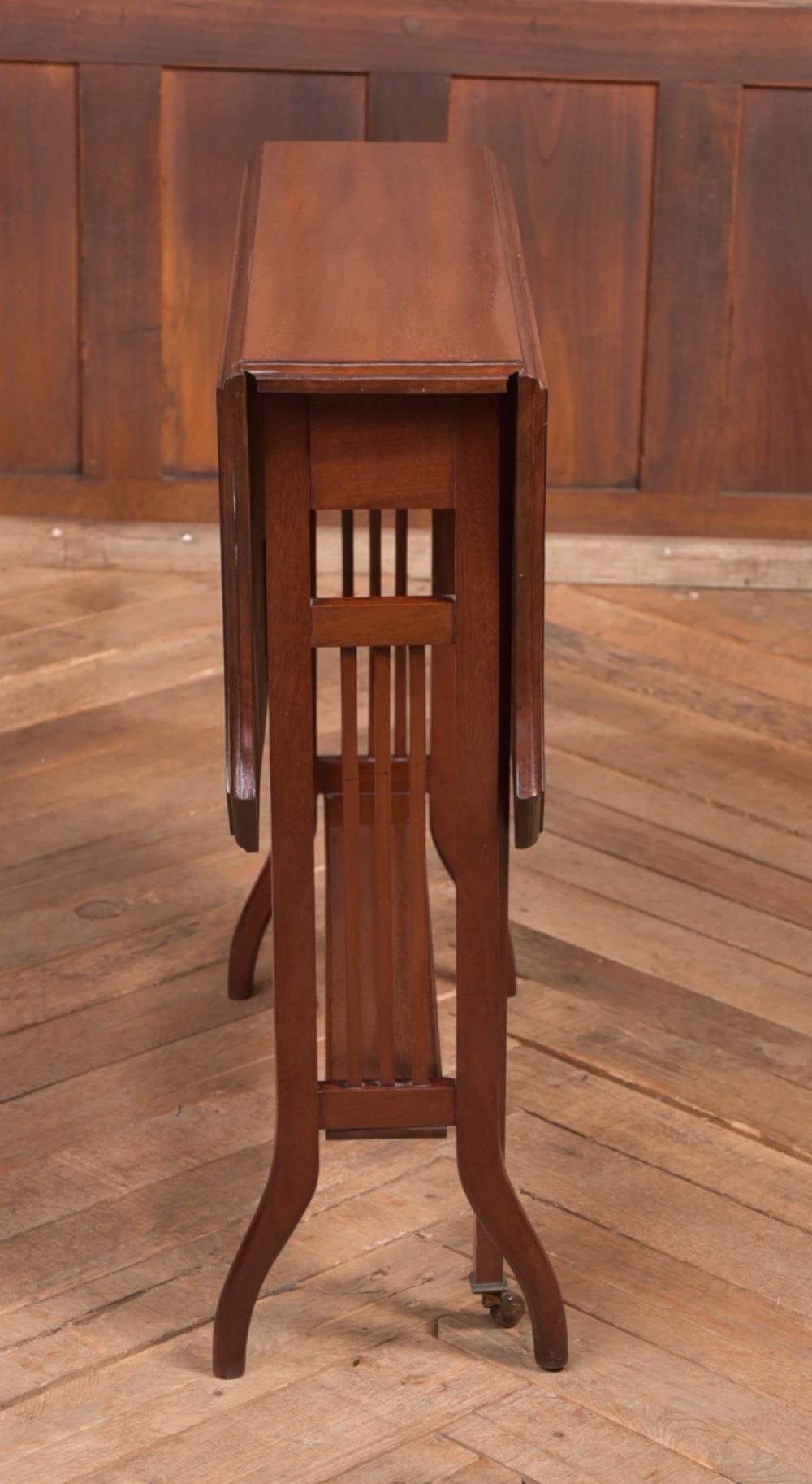 An Edwardian mahogany Sutherland side table shown in a folded state with deep drop leaves, slatted end supports, and curved legs with brass castors. View VI