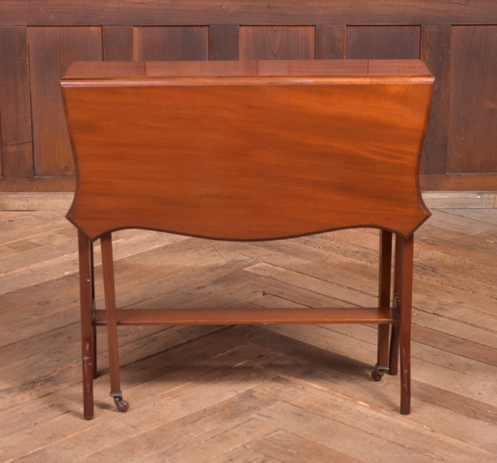 An Edwardian mahogany Sutherland side table shown in a folded state with deep drop leaves, slatted end supports, and curved legs with brass castors. View VII