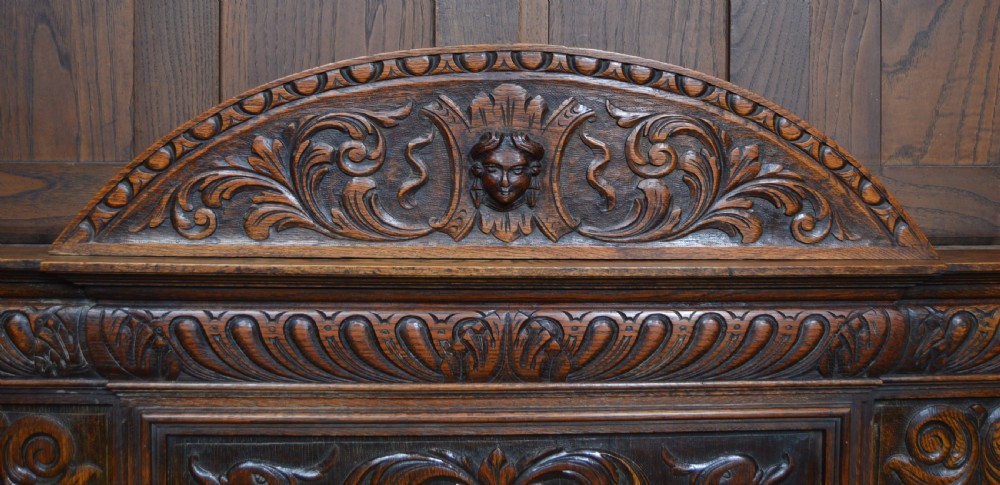 A heavily carved dark oak monks bench featuring Green Man foliate face motifs, ornate scrollwork, a raised back panel, and a hinged lid seat. View II