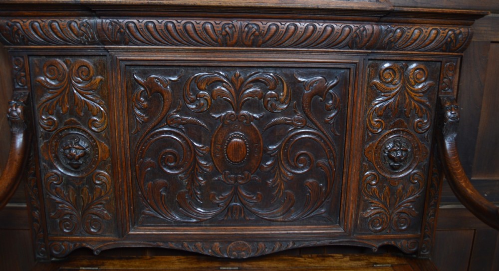 A heavily carved dark oak monks bench featuring Green Man foliate face motifs, ornate scrollwork, a raised back panel, and a hinged lid seat. View V