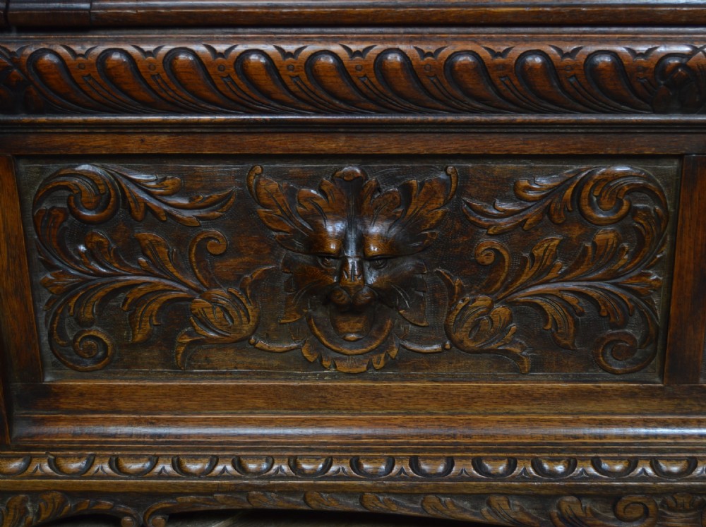 A heavily carved dark oak monks bench featuring Green Man foliate face motifs, ornate scrollwork, a raised back panel, and a hinged lid seat. View VI