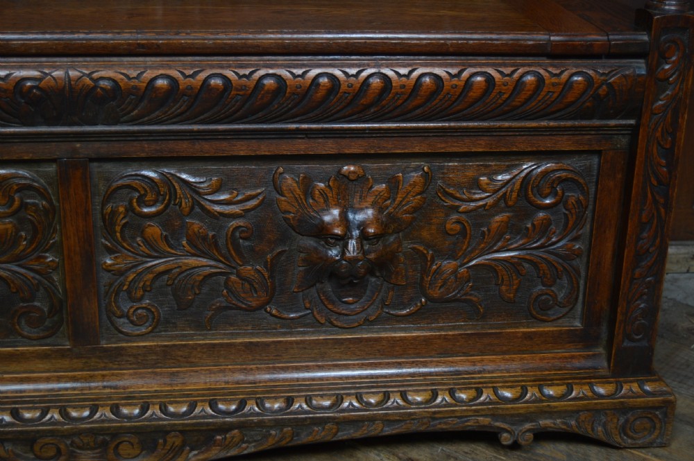 A heavily carved dark oak monks bench featuring Green Man foliate face motifs, ornate scrollwork, a raised back panel, and a hinged lid seat. View VII