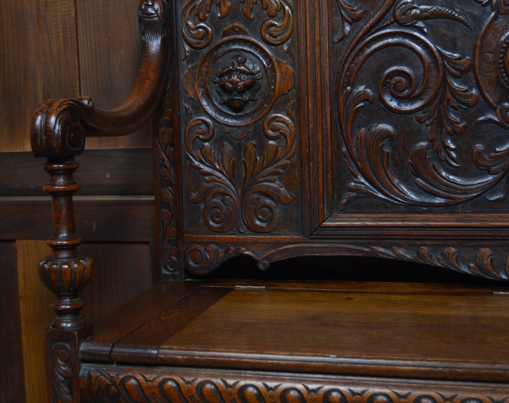A heavily carved dark oak monks bench featuring Green Man foliate face motifs, ornate scrollwork, a raised back panel, and a hinged lid seat. View IX