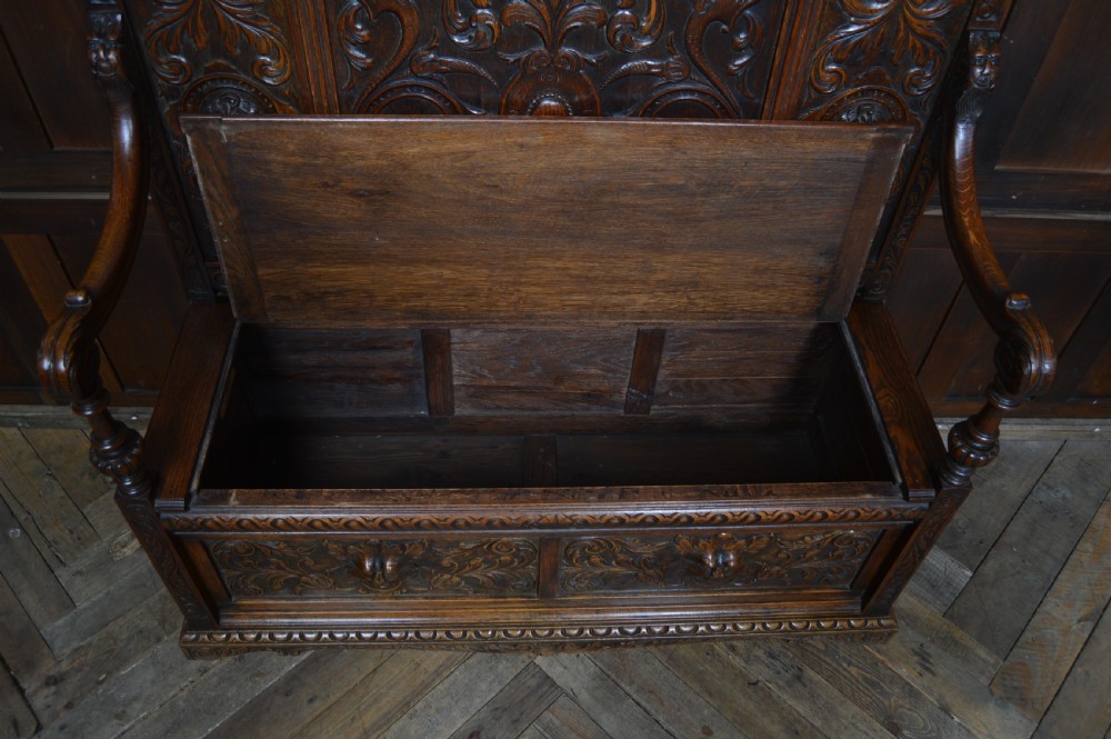 A heavily carved dark oak monks bench featuring Green Man foliate face motifs, ornate scrollwork, a raised back panel, and a hinged lid seat. View XI