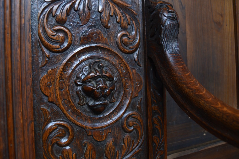 A heavily carved dark oak monks bench featuring Green Man foliate face motifs, ornate scrollwork, a raised back panel, and a hinged lid seat. View XIV