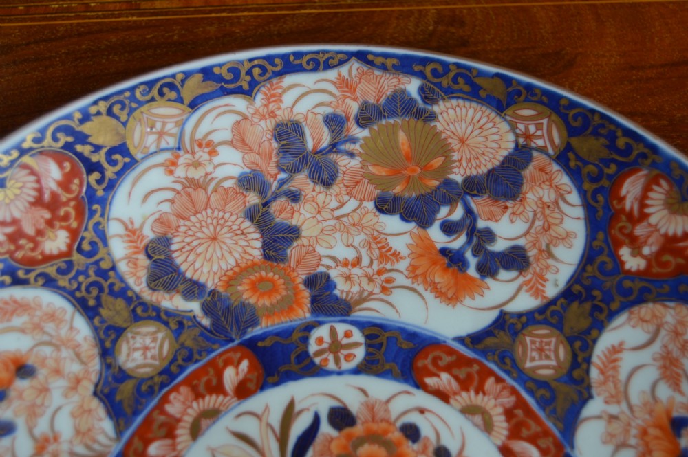 A circular Japanese Imari porcelain plate decorated with hand-painted cobalt blue, rust red, and gold floral patterns surrounding a central floral vase motif. View III