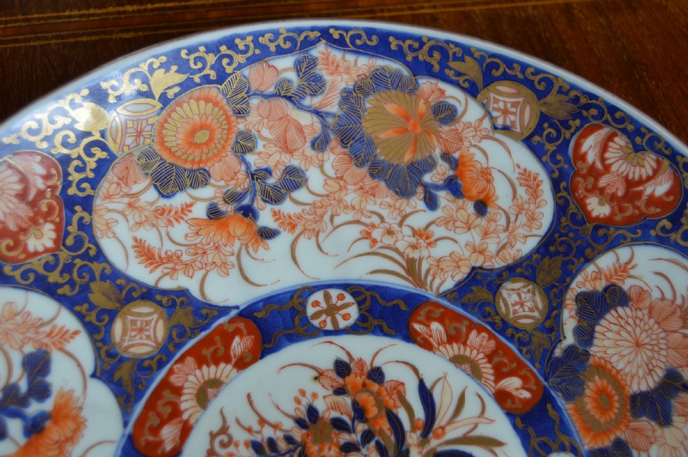 A circular Japanese Imari porcelain plate decorated with hand-painted cobalt blue, rust red, and gold floral patterns surrounding a central floral vase motif. View V