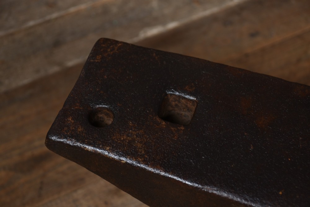 A dark brown cast iron blacksmith anvil featuring a flat face, conical horn, and square hardy hole resting on a wooden floor. View VII