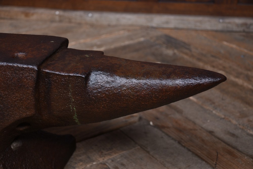 A dark brown cast iron blacksmith anvil featuring a flat face, conical horn, and square hardy hole resting on a wooden floor. View VIII
