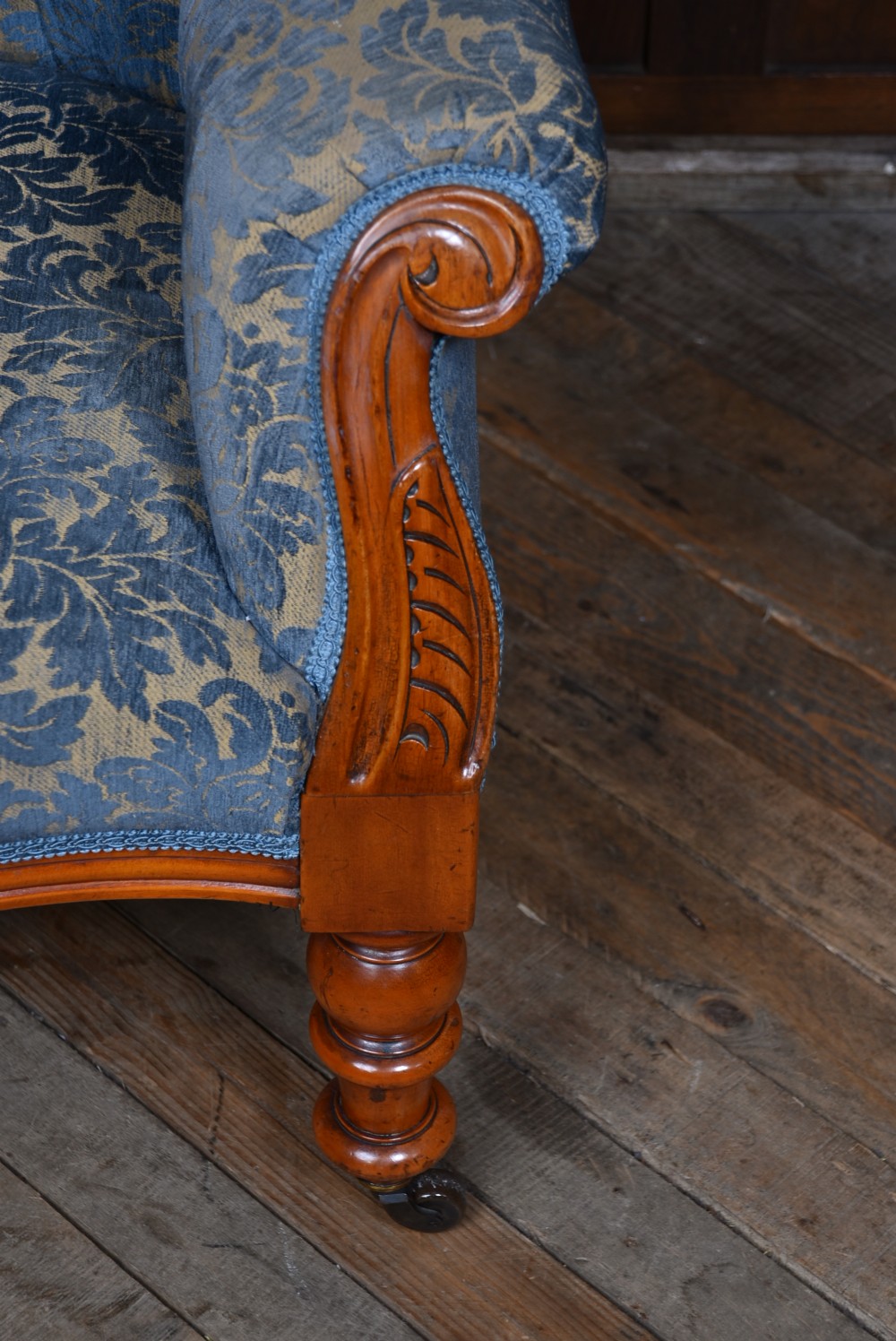 A Victorian mahogany armchair with a carved top cresting, deep buttoned blue patterned fabric upholstery, padded scroll arms, and turned front legs on brass castors. View V