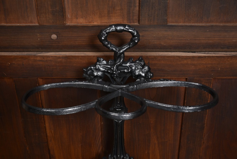 An ornate black cast iron umbrella stand featuring elaborate scrollwork, dual holding rings, a looped handle, and a contoured base tray. View VI