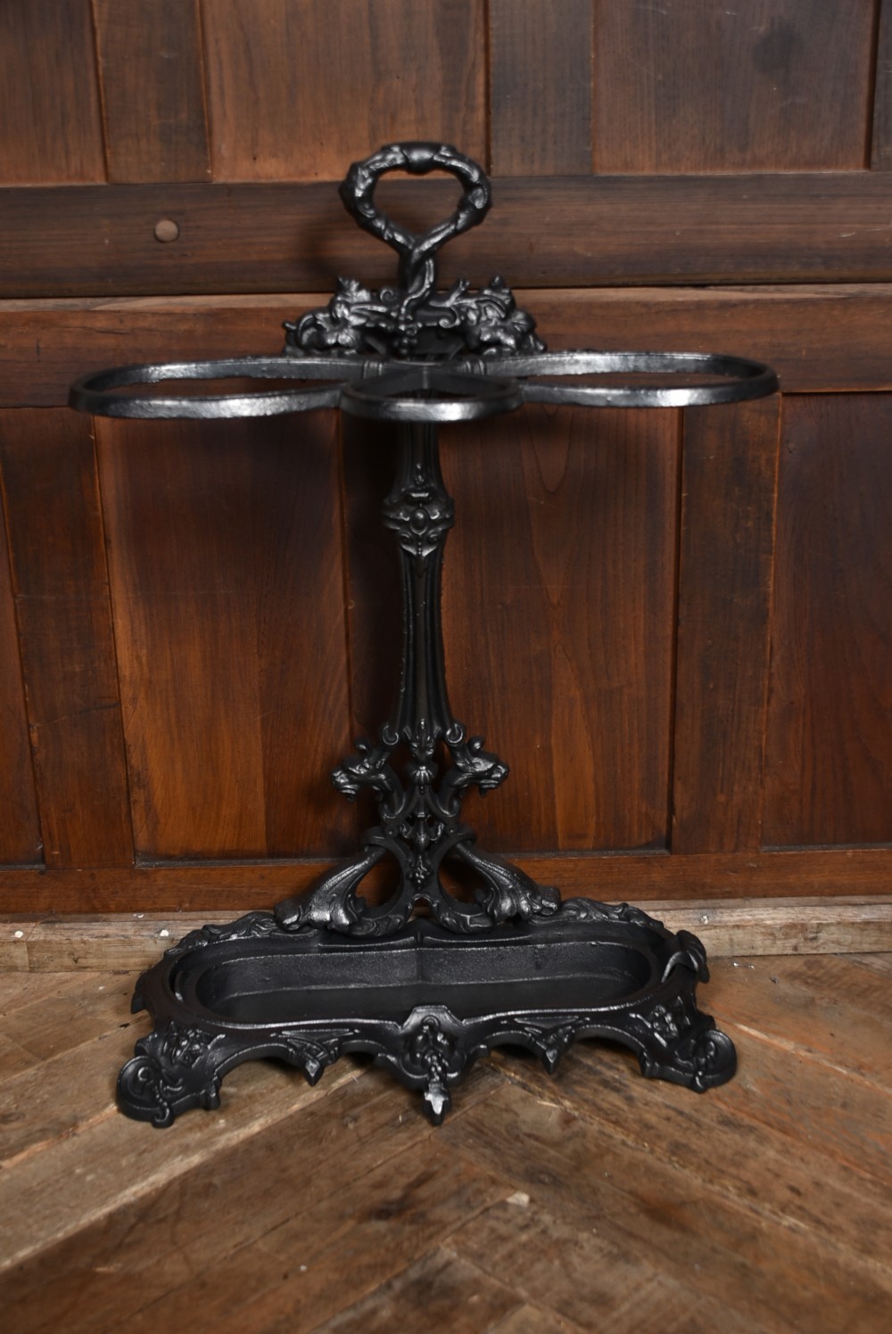 An ornate black cast iron umbrella stand featuring elaborate scrollwork, dual holding rings, a looped handle, and a contoured base tray. View VIII