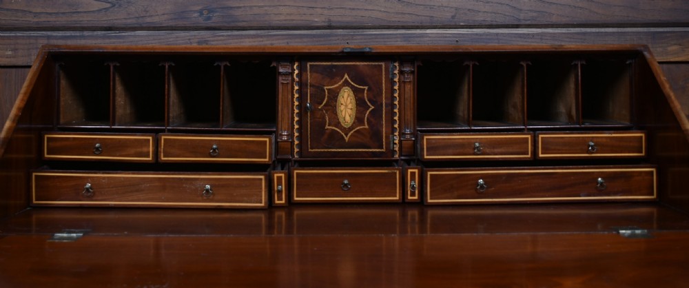 An antique Georgian mahogany slant-front writing bureau featuring four graduated drawers with brass handles and central oval marquetry inlay on the fall flap. View X