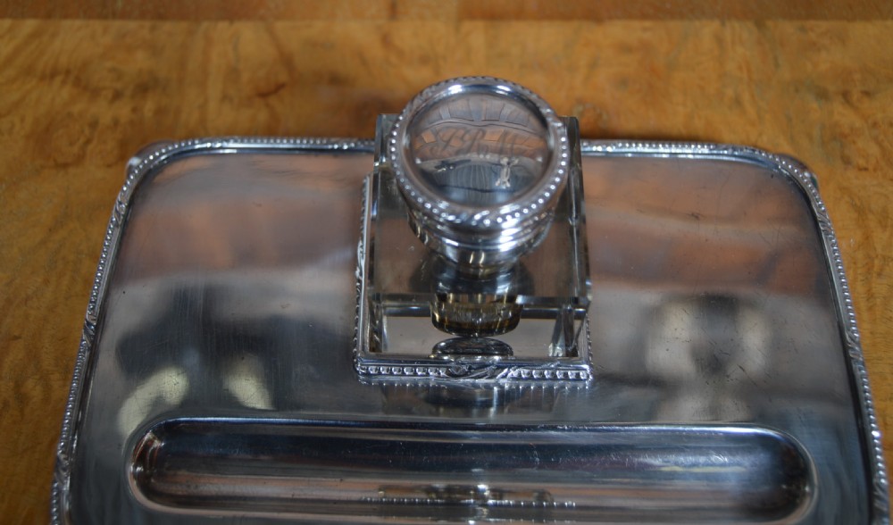 An electroplated nickel silver inkstand featuring a rectangular tray with a built-in pen trough, raised on ball feet, holding a central square glass inkwell with a silver lid. View V