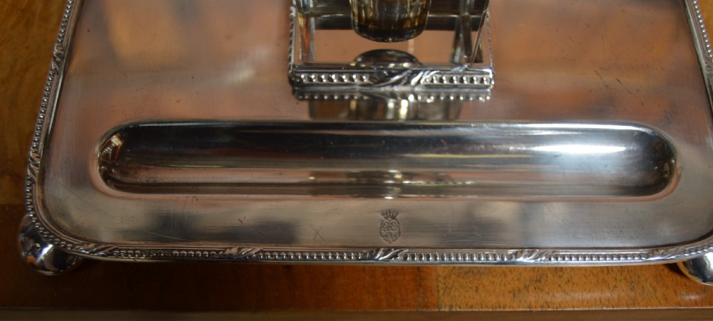 An electroplated nickel silver inkstand featuring a rectangular tray with a built-in pen trough, raised on ball feet, holding a central square glass inkwell with a silver lid. View VII