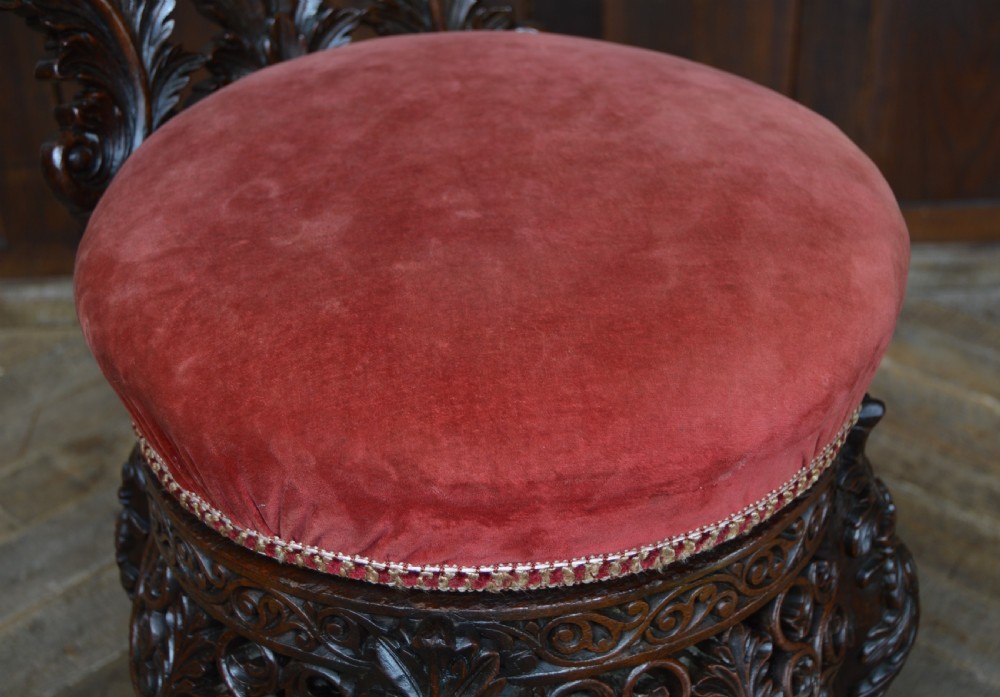 An antique Anglo-Burmese carved padouk wood music chair featuring an intricate openwork back, round crimson velvet upholstered seat, and carved legs. View III