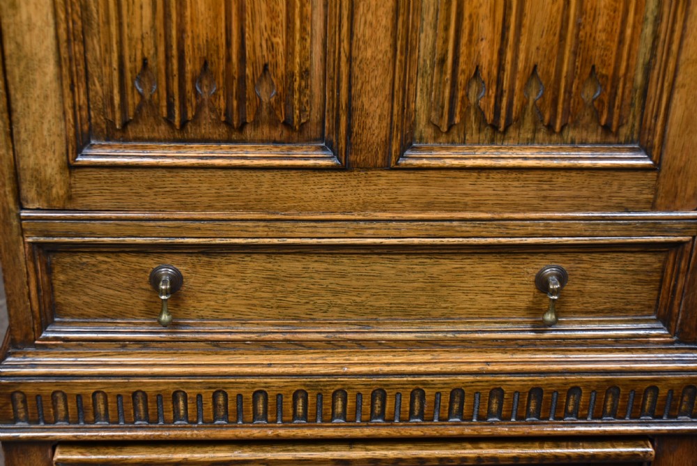 An Edwardian oak writing bureau featuring carved linenfold panel doors, a drawer with brass drop handles, and turned legs. View V