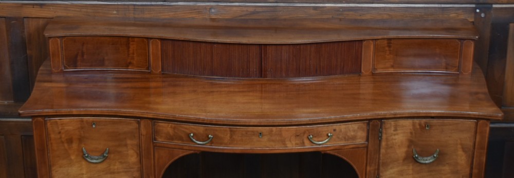 An Edwardian mahogany sideboard featuring satinwood string inlay, a curved central drawer, flanking cupboard doors, a raised superstructure, and square tapered legs. View VI