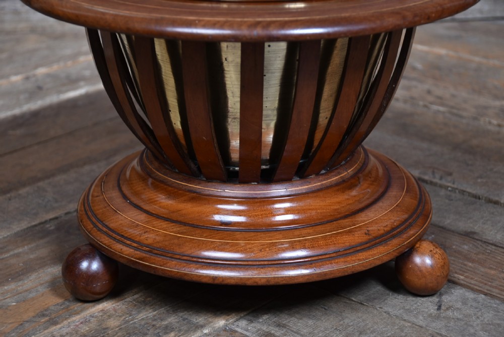 A circular mahogany planter with vertical open slats, turned ball feet, a brass swing handle, and a removable brass liner. View II
