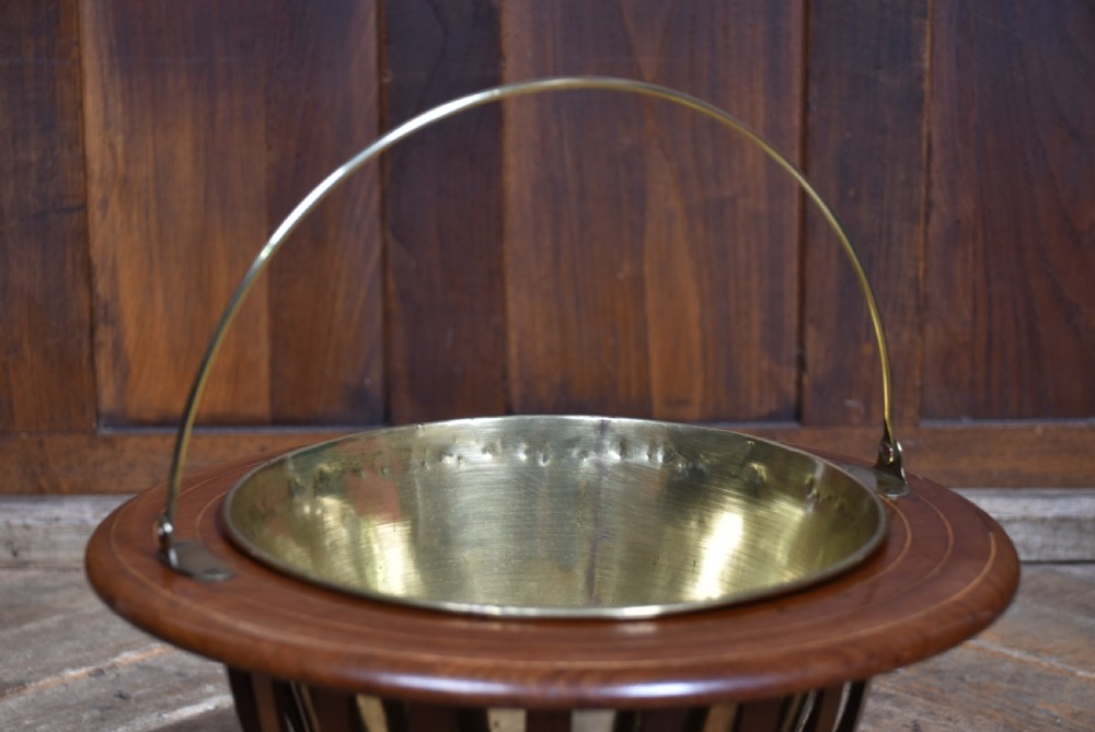 A circular mahogany planter with vertical open slats, turned ball feet, a brass swing handle, and a removable brass liner. View V