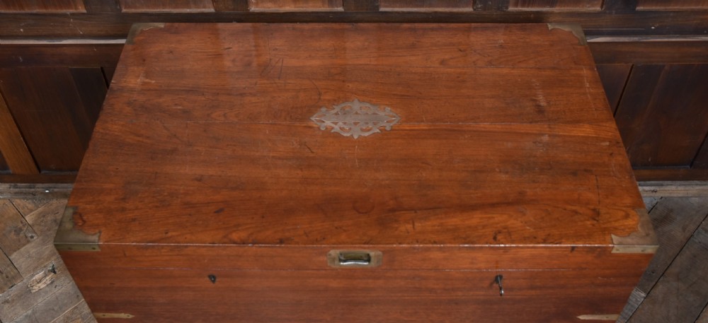 A nineteenth-century rectangular teak coffer with flush brass corner brackets, brass escutcheons, and an inlaid top cartouche, raised on a wooden stand with square legs. View VI