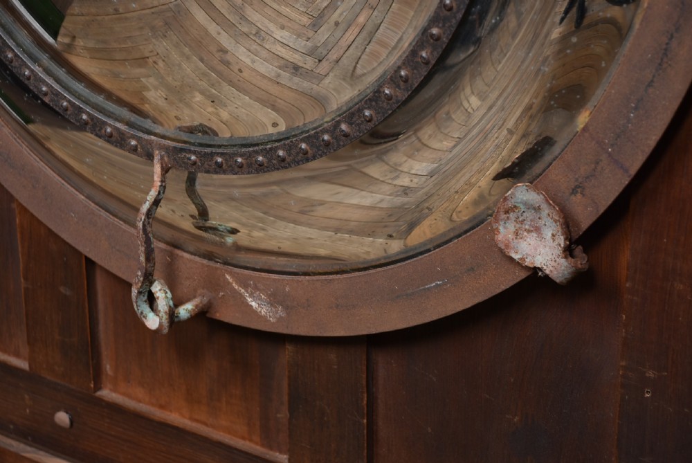A circular convex wall mirror featuring a patinated dark metal frame, riveted inner rim, forged scroll mounts, and foxed glass reflection. View IX