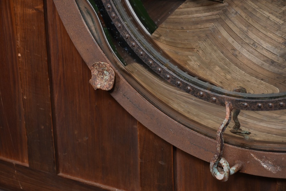 A circular convex wall mirror featuring a patinated dark metal frame, riveted inner rim, forged scroll mounts, and foxed glass reflection. View X
